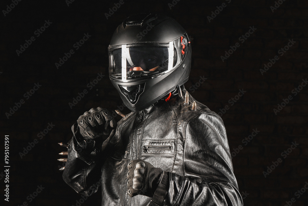 Brutal motorcyclist rider in black helmet and black studded leather jacket on a dark background.