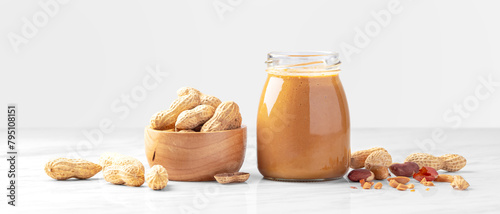 Peanut butter in jar and roasted peanuts in wooden bowl on white marble table