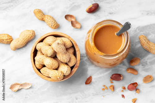 Peanut butter in glass jar with spoon and peanuts in wooden bowl on white marble table