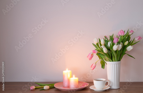 cup of coffee and spring tulips in vase with burning candles on wooden shelf
