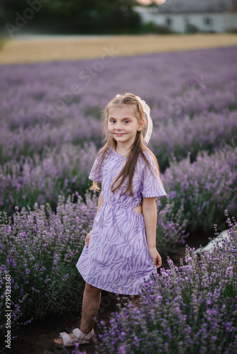 A smiling girl in a purple dress walks among lavender flowers with sunlight on a summer day. A child girl goes into a lavender field at sunset. Kid looking at the camera.