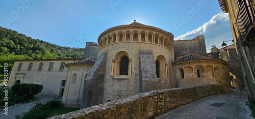 Fotografia Old, beautiful stone church buildings in the south of France with a cobbled road, circular walls and a cross under a bright blue sky with green trees in the background