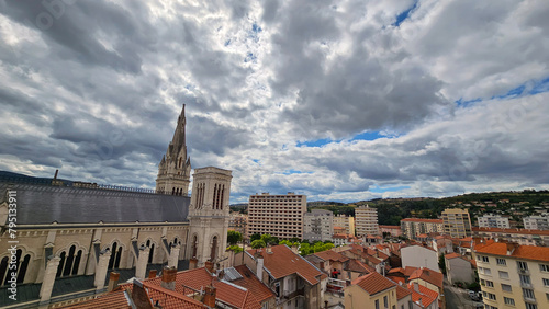 View of Saint-Chamond in the Loire Department, France with roof tops, apartments and church under a cloudy sky; Église Notre-Dame de Saint-Chamond, surrounded by flats, with hills in the distance