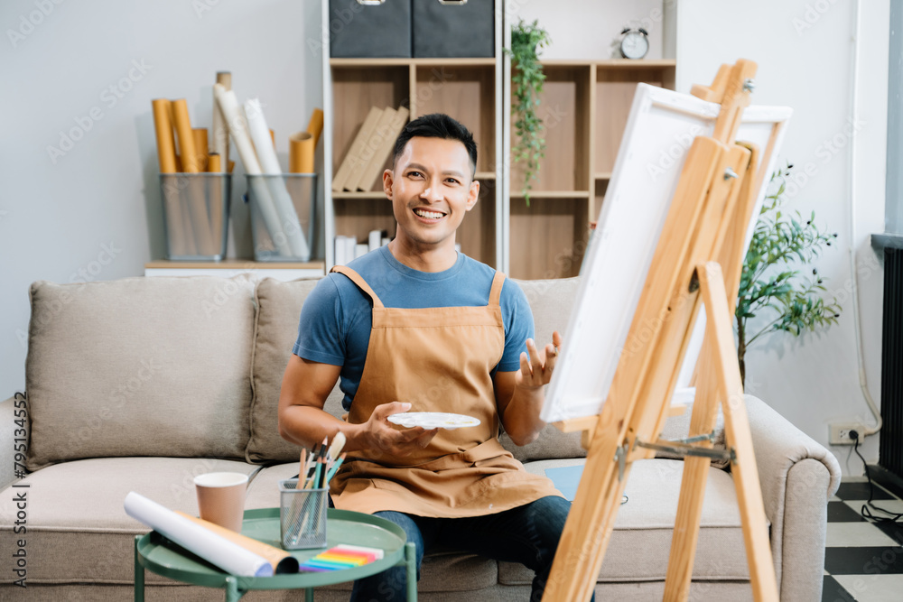 Asian male painter do artwork in art workshop, painting supplies, oil pastels, two canvas easel, creative space with paintbrush in art studio