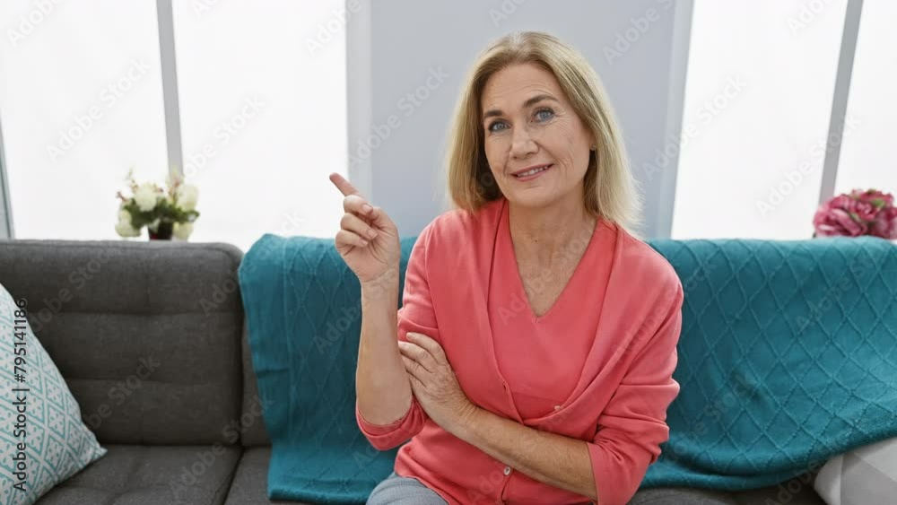 Radiant middle-aged blonde woman smiling cheerfully on sofa, pointing with a joyous expression at something off-side within her cozy indoor living room. Radiant middle-aged blonde woman smiling cheerfully on sofa, pointing with a joyous expression at something off-side within her cozy indoor living room.