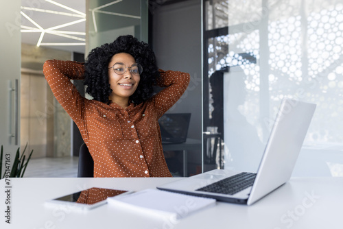 A young Black businesswoman enjoys a moment of relaxation at her desk inside a modern office. She smiles, leaning back with hands behind her head, near her laptop. photo