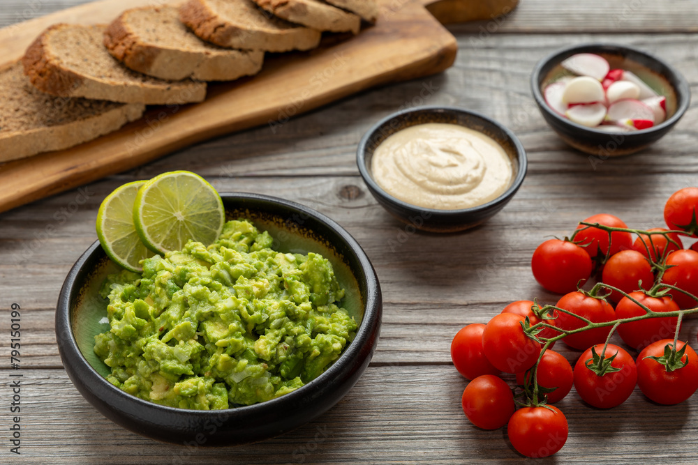 Fresh chunky guacamole decorated with slices of lime, homemade bread slices on a wooden kitchen board, cherry tomatoes, tahini sauce, and red radishes on the table. Homemade, healthy vegan meal. Fresh chunky guacamole decorated with slices of lime, homemade bread slices on a wooden kitchen board, cherry tomatoes, tahini sauce, and red radishes on the table. Homemade, healthy vegan meal.