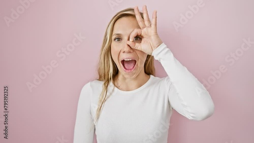 Blonde bombshell radiates shock and surprise! gutsy young woman makes a cheeky 'ok' gesture, peeks through fingers over an isolated pink wall background. photo