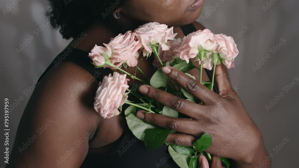 Studio portrait of young tender African American woman holding bouquet of flowers to her chest, touching roses lovingly and posing for camera