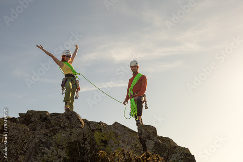 Two people are standing on a mountain, one of them is wearing a yellow shirt. They are both wearing helmets and harnesses, and they are holding onto a rope. Scene is adventurous and exciting