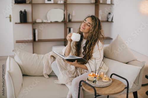 Young woman reading book and drinking tea, sitting on sofa in a cozy living room.