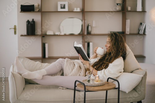 Young woman reading book and drinking tea, sitting on sofa in a cozy living room.