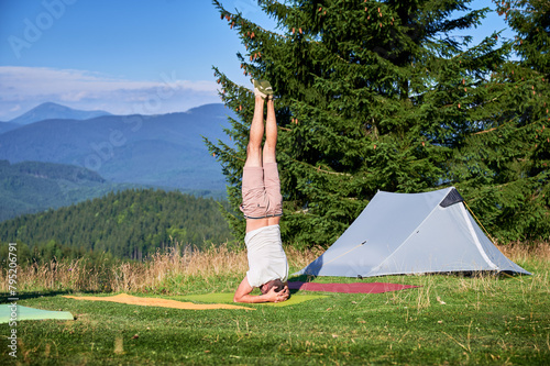 Man performing headstand, yoga pose Sirsasana on a yoga mat in a scenic outdoor setting. Person in grassy area, gray camping tent on background, under clear blue sky.