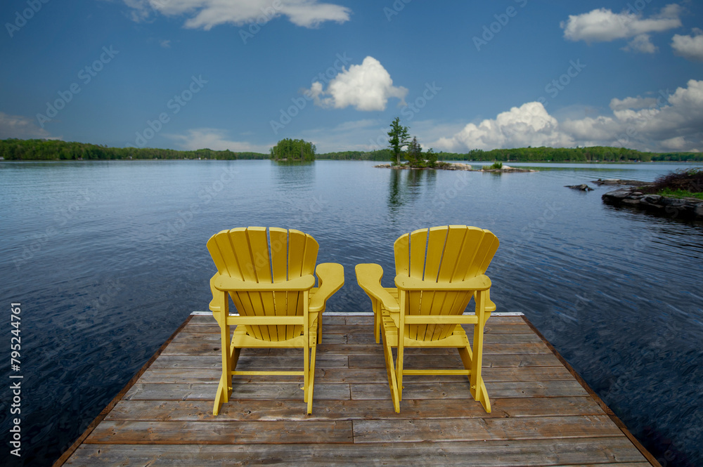 Two wooden Adirondack chairs on a dock greet the tranquil Muskoka morning, overlooking the serene blue lake waters. Across the way, cottages nestled among green trees complete the picturesque scene. Two wooden Adirondack chairs on a dock greet the tranquil Muskoka morning, overlooking the serene blue lake waters. Across the way, cottages nestled among green trees complete the picturesque scene.