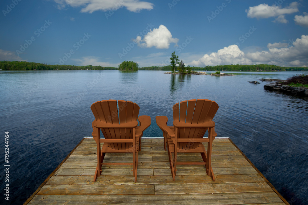 Two yellow Adirondack chairs on a wooden dock in Muskoka, overlooking the tranquil blue lake waters. Across, cottages nestled among green trees complete the scene.