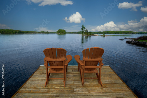 Two yellow Adirondack chairs on a wooden dock in Muskoka  overlooking the tranquil blue lake waters. Across  cottages nestled among green trees complete the scene.