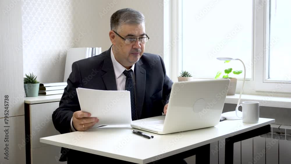 A man in a suit and tie works in the office. He looks through business papers and types on the computer. Suitable for content about working in the office, running a business or remote work. A man in a suit and tie works in the office. He looks through business papers and types on the computer. Suitable for content about working in the office, running a business or remote work.
