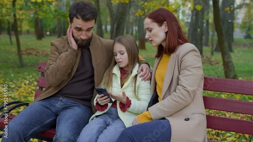 Caucasian family at city bench together outdoors autumn sad upset confused worried with mobile phone bad news smartphone failure little daughter girl child kid with parents mother woman father man