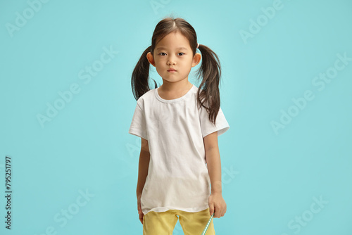 Asian ethnicity little five years old girl, wearing in white t shirt and colorful yellow jeans, has hair is gathered on the sides in a ponytail standing over blue background with a free copy space.