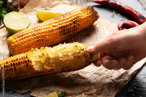 Slika na platnu Caucasian hand holds half eaten grilled corn on cob with lime and other spices o