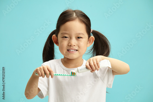 Charming little asian ethnicity child girl squeezes toothpaste out of a tube onto a toothbrush, smiles happily, wears a casual white T-shirt, stands on a blue isolated background.