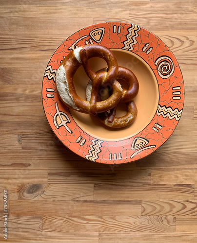 Two pretzels lie together in one large deep round empty orange plate with ethnic ornaments on a wooden background.