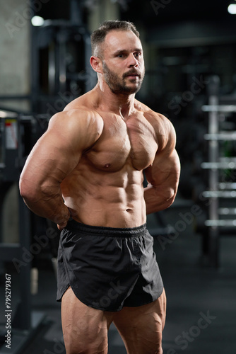 Attractive bearded bodybuilder performing healthy, strong body in gym. Portrait of Caucasian male weightlifter in black shorts, demonstrating muscles, with hands behind back. Concept of bodybuilding.