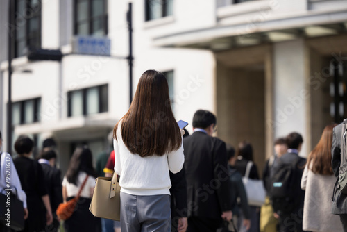 ビジネス街の横断歩道を渡りながらスマホを見る日本人女性の後ろ姿