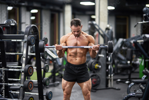 Strong male athlete holding barbell, weight lifting in gym. Front view of muscular Caucasian man lifting barbell equipment, exercising, against blurred background. Bodybuilding, weightlifting concept.