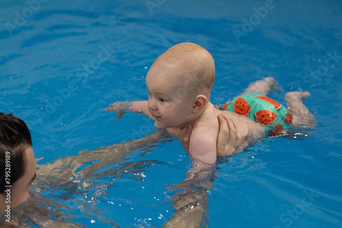 Smiling baby in the fun swim panties swimming with father in swimming pool. Swimming lessons for newborn.
