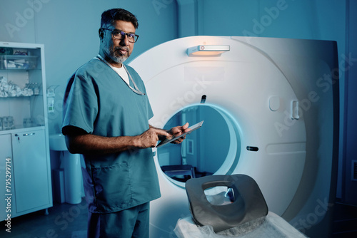 Medium long shot of biracial doctor standing by MRI scanner holding digital tablet looking at camera, copy space