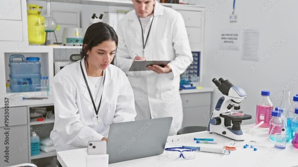 A man and woman in white lab coats working together in a science laboratory filled with research equipment A man and woman in white lab coats working together in a science laboratory filled with research equipment
