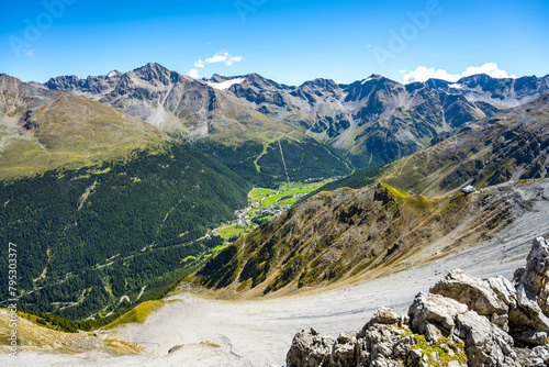 A breathtaking summer panorama of the Sulden Valley in Italy, showcasing the lush green valley contrasted with the rugged mountain peaks under a blue sky.