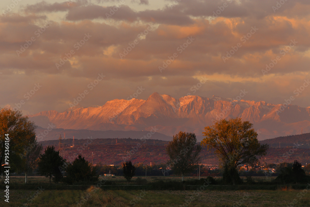 primo piano di parte della catena montuosa delle Alpi Giulie, sotto un cielo nuvoloso, colorata di arancione dalla luce del sole al tramonto, vista in lontananza dalla campagna distante