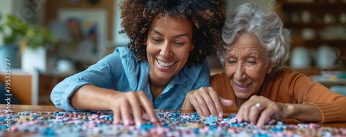 A smiling female caregiver aids a senior woman with a puzzle at home, depicting companionship and assistance. © vadymstock