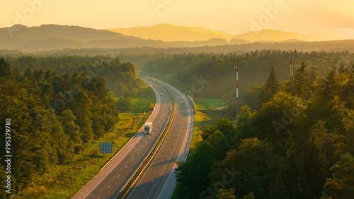 AERIAL: Trucks and cars travel along the scenic highway in golden evening light. Asphalt motorway winds through the beautiful wooded and hilly countryside, bathed in the rays of setting summer sun.