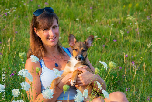 PORTRAIT, DOF: Blooming meadow with a pretty lady posing with her young puppy. Smiling woman lovingly holds her small puppy amidst a field of wildflowers. Moments of happiness with a furry companion.