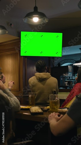 Vertical shot of friends sit in sports pub and watch live soccer match broadcast on TV. Fans gathered to cheer and support team playing in tournament. Player scores a goal and crowd celebrate.