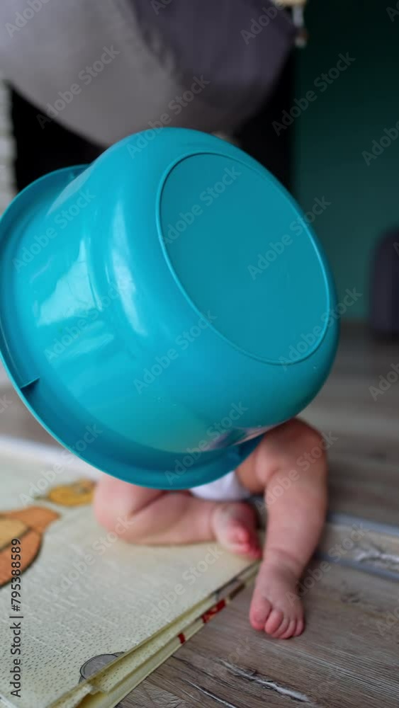 Little infant kid sits on the floor covered with blue washtub. Blond baby drops the wash bowl. Vertical video.