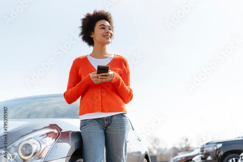 Happy African American woman with curly hair holding mobile phone, standing near car, looking away © Maria Vitkovska