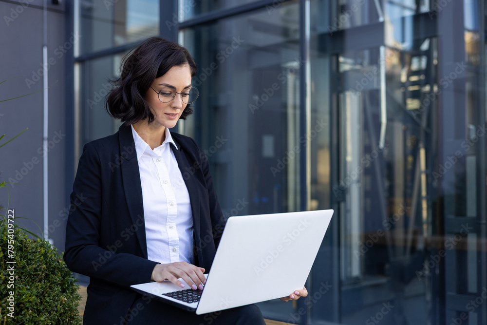 A smiling young woman in a business suit is sitting outside an office building, working and using a laptop A smiling young woman in a business suit is sitting outside an office building, working and using a laptop