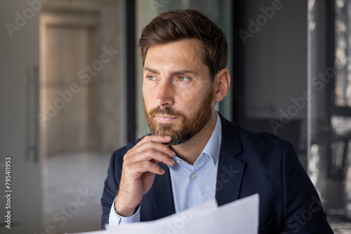 Close-up photo of a young businessman sitting in a modern office in a suit, holding papers in his hands and seriously thinking about solving a problem and a deal