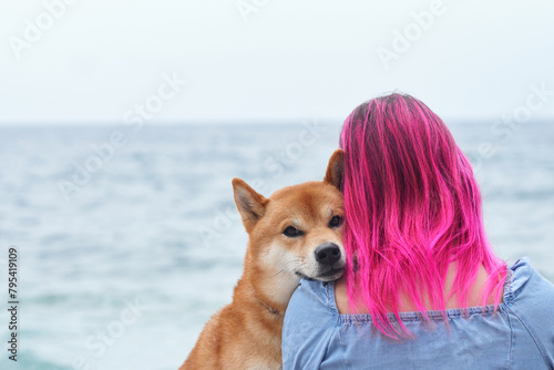 A Shiba Inu dog cozily nestles into a woman with striking pink hair, overlooking the tranquil sea. Their affectionate embrace embodies a special human-animal bond