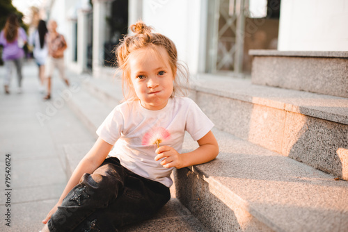 Funny child girl 4-5 year old wearing casual clothes sitting on stairs over city street at background outdoor. Looking at camera.