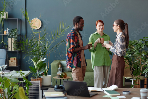 Creative business team of three people communicating standing in modern office at coffee break against blue wall copy space