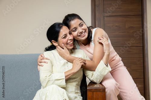 Indian Mother and Teen young daughter sitting on sofa at home looking towards the camera ,Happy Young teen hug Mother in living room having candid moments