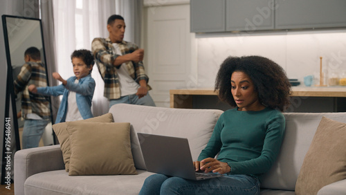Quarantine family problems African American woman female mom trying to work online laptop pc sit sofa living room while loud man male dancing dad little boy child kid son distract mother businesswoman © Yuliia