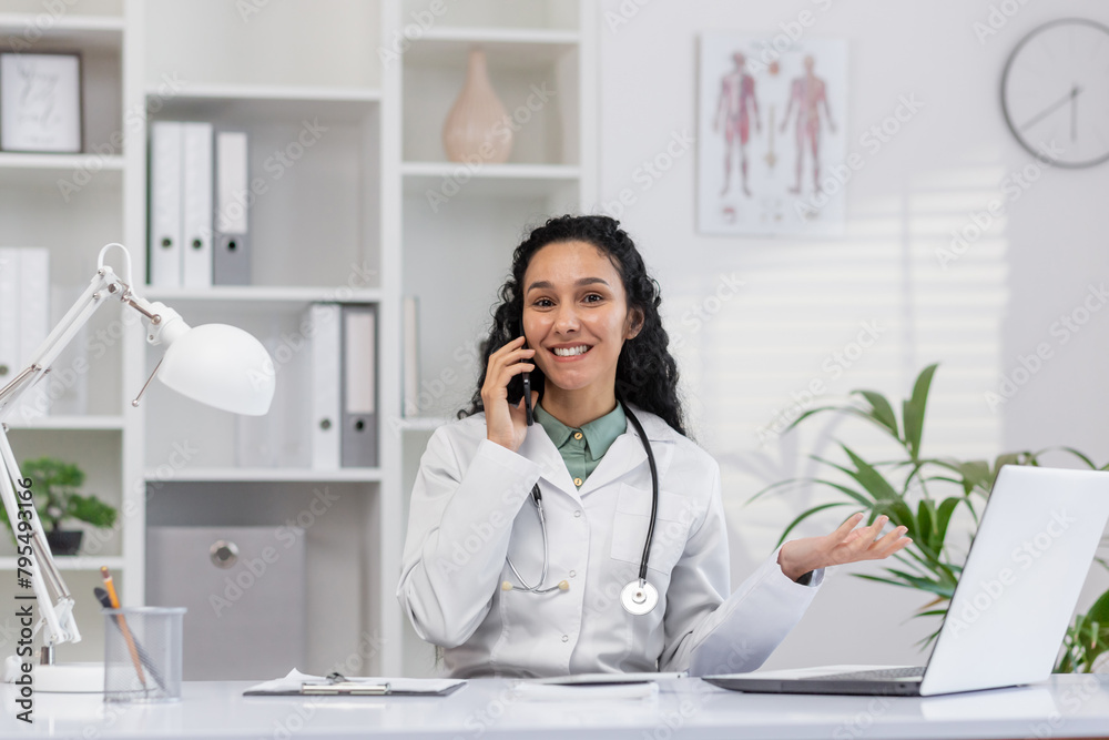 Confident Hispanic female doctor in white lab coat consulting over the phone, smiling in a well-lit, modern office with medical posters and plants. Confident Hispanic female doctor in white lab coat consulting over the phone, smiling in a well-lit, modern office with medical posters and plants.