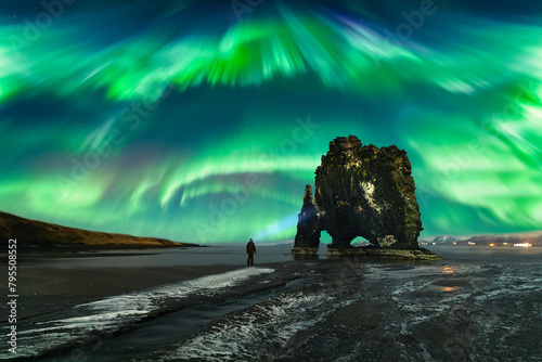 A man with a lantern (headlight) watches the  Northern Lights (Aurora Borealis) dance across the night sky next to the famous volcanic rock formation Hvítserkur. Iceland photo