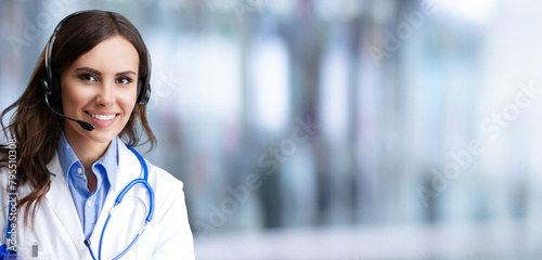 Portrait image of happy smiling female doctor in headset, against blurred modern clinic office background, blank mock empty area for ad slogan text. Medical call center. Wide banner
